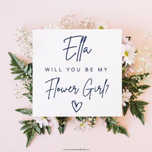 Pode incluir: Um cart&atilde;o branco com o texto "Ella, voc&ecirc; quer ser minha dama de honra?" em escrita cursiva azul marinho com um pequeno cora&ccedil;&atilde;o desenhado abaixo do texto. O cart&atilde;o est&aacute; rodeado de flores brancas e folhas verdes sobre um fundo rosa claro.