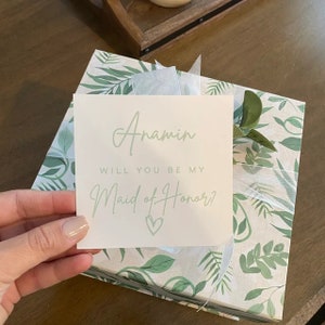 Pode incluir: Um cart&atilde;o branco com texto verde que diz "Anamin, voc&ecirc; quer ser minha dama de honra?" com um cora&ccedil;&atilde;o verde. O cart&atilde;o est&aacute; em cima de uma caixa de presente com um padr&atilde;o de folhas tropicais verdes e brancas.