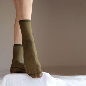 May include: A pair of sheer green socks with a ribbed top. The socks are worn on a person's feet and are shown against a light brown background.