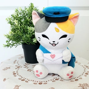 May include: A white plush cat with gray and yellow markings wearing a blue hat with a black brim. The cat is holding a white envelope with a red heart on it. The cat is sitting on a white table with a brown floral pattern.