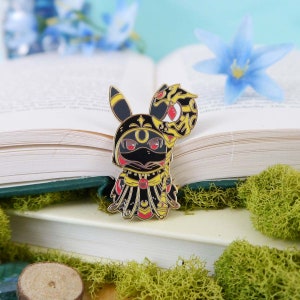 May include: A gold-colored enamel pin featuring a cartoon character with a black and yellow costume, a red and yellow snake tail, and a black mask. The character is wearing a gold and red robe with a crescent moon design.