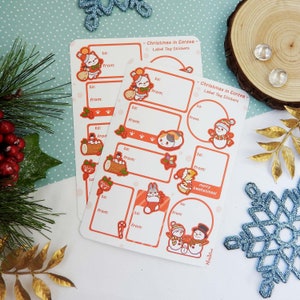 Könnte beinhalten: Zwei Bögen mit Weihnachtsetiketten-Aufklebern mit verschiedenen festlichen Illustrationen, darunter eine Katze, ein Schneemann und ein Hase. Jeder Aufkleber hat einen rechteckigen Bereich für "an" und "von" Details. Der Text "Christmas in Eorzea - Label Tag Stickers" ist oben aufgedruckt.