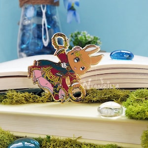 May include: Gold-tone enamel pin featuring a cartoon rabbit wearing a red and gold outfit, holding a golden rope. The rabbit has brown fur and a pink nose. The pin is on a book with a green moss background.
