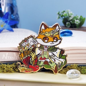Könnte beinhalten: Goldfarbene Emaille-Anstecknadel mit einem Fuchs, der eine Krone und eine grün-weiße Robe trägt, ein Buch und eine Feder hält. Der Fuchs steht auf einem rot-goldenen Vogel mit langem Schwanz.