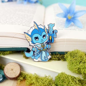 May include: A blue and white enamel pin featuring a cartoon character with a crown and a staff. The character has a blue tail and fins, and is holding a staff with a blue star on top. The pin is set against a background of a book and moss.