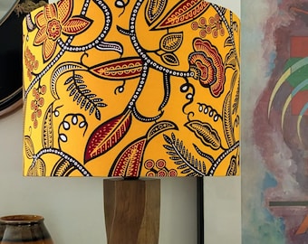 May include: A yellow lampshade with a floral pattern in red, black, and white sits atop a wooden lamp base. The lamp is on a dark surface, with a vase and artwork in the background. The lampshade's design features stylized flowers and leaves.