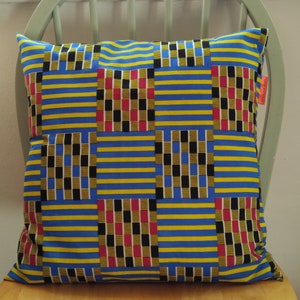 May include: A blue and yellow striped cushion with a geometric pattern in black, red and white. The cushion has a label that says "Studio & Co."