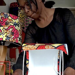 May include: A person working on a lampshade. The lampshade has a vibrant pattern of red, yellow, and black geometric shapes. The person is wearing a black top and a pink headband. Other lampshades with similar patterns are in the background.