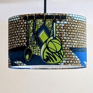 May include: A cylindrical lampshade with a white interior and a colourful exterior. The exterior features a blue, green and brown pattern of kitchen utensils hanging on a rack.