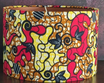 May include: Cylindrical lampshade with a vibrant African wax print. The fabric features a repeating pattern of red, yellow, and brown organic shapes, accented with black floral motifs. The interior is a warm, neutral tone.