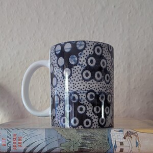 May include: A white ceramic mug featuring a blue and white circular pattern. The mug has a white handle and is placed on a book titled "Boy Akosua Abes". The mug is suitable for tea or coffee and is a standard size.