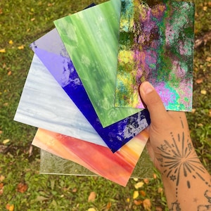 May include: A collection of rectangular stained glass sheets in various colors including iridescent purple and green, solid green, deep blue, light blue, and a peach and yellow blend, fanned out to display their distinct textures and hues.