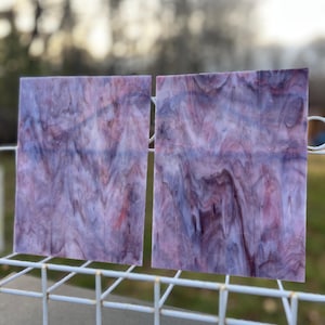 May include: Two square pieces of stained glass with a marbled effect. The glass features shades of purple, red, and white, creating a swirling pattern. The glass is held by a white wire rack, with a blurred outdoor background.