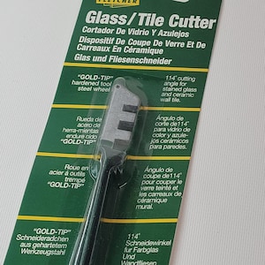 May include: A green and white packaging for a glass and tile cutter. The cutter has a black handle and a silver wheel. The packaging includes the text "Glass/Tile Cutter" and "GOLD-TIP".