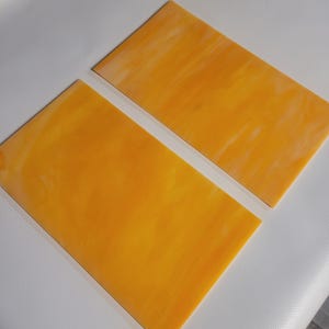May include: Two rectangular sheets of stained glass in shades of yellow and orange. The glass has a marbled appearance with lighter streaks throughout. The sheets are positioned diagonally, with one slightly overlapping the other.