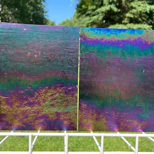 May include: Two iridescent glass panels with a vibrant display of colors, including purple, green, and blue. The panels are positioned on a white metal rack, with a backdrop of green foliage and a blue sky.