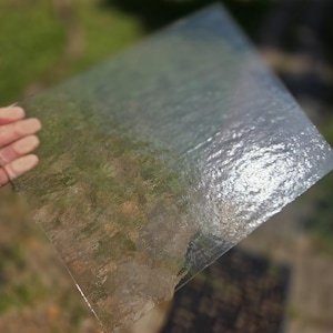 May include: A square sheet of clear, textured glass with a rippled, uneven surface that creates a distorted, translucent effect. The glass is held at an angle, allowing light to reflect off the irregular ridges and bumps across its entire surface.