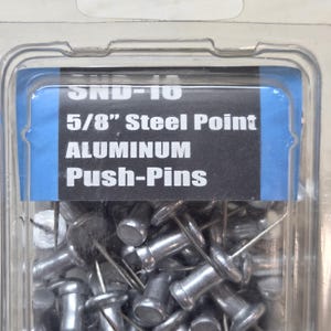 May include: A package of aluminum push pins with steel points. The label reads "SND-10 5/8" Steel Point ALUMINUM Push-Pins". The push pins are silver and have a round head and a sharp point. The package is clear plastic.