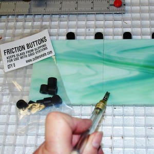 May include: A close-up of a glass cutter scoring a piece of green and white marbled glass. The glass is sitting on a grid cutting mat. A bag of black friction buttons is in the background. The bag reads "Friction Buttons Keeps Glass From Slipping For Use With Grid Systems QTY 8 www.cuttersmate.com"