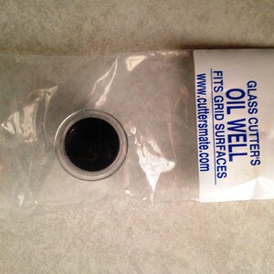 May include: A small, black, round oil well for a glass cutter. The oil well is packaged in a clear plastic bag with a white label that reads "GLASS CUTTER'S OIL WELL FITS GRID SURFACES www.cuttersmate.com".