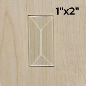 May include: A 1" x 2" wooden plaque with a raised, geometric design. The design is a rectangle with a vertical line down the center and two diagonal lines extending from the center line to the corners of the rectangle.