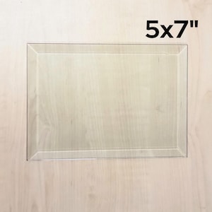 May include: A clear glass picture frame with a 5x7 inch opening. The frame is on a light wood surface.