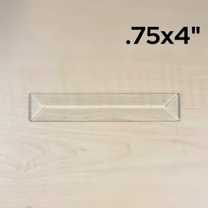 May include: A clear glass prism measuring .75 inches by 4 inches. The prism is on a light wood surface.