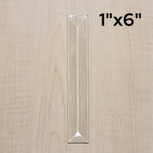 May include: A clear glass prism with beveled edges, measuring 1 inch by 6 inches. The prism is on a light wood surface.
