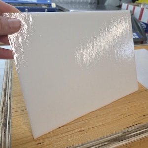 May include: A rectangular, off-white, glossy tile is held by a hand. The tile has a smooth, reflective surface. It sits on a light-colored wooden surface. The tile appears to be new and unused.