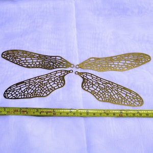 May include: Four gold-colored metal dragonfly wings with intricate cut-out designs. The wings are arranged in a cross pattern and measure approximately 10 inches in length.