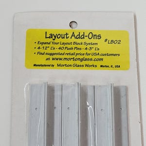 May include: Layout Add-Ons package with silver metal pieces. The yellow label reads "Layout Add-Ons" and includes text about the product and the manufacturer, Morton Glass Works, in Morton, IL, USA. The package includes 4-12" L's and 40 push pins.