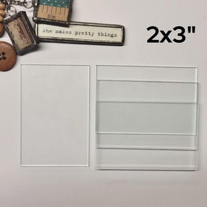 May include: Two pieces of clear glass, one measuring 2x3 inches, the other measuring 3x4 inches. The glass is smooth and has a slight sheen. The text "she makes pretty things" is visible on a small metal sign in the background.