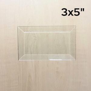 May include: A clear, rectangular acrylic display case, measuring 3x5 inches, is shown against a light wood-grain background. The case has a simple, clean design, suitable for showcasing small items.