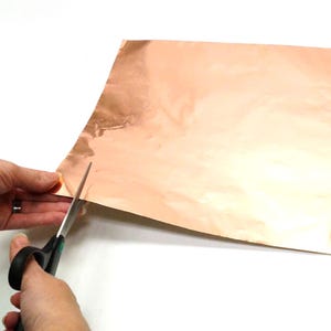 May include: A person is cutting a sheet of copper foil with scissors. The foil is a shiny, metallic copper color.