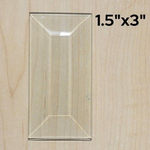 May include: A clear glass tile with a beveled edge, measuring 1.5 inches by 3 inches.