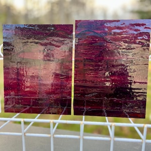 May include: Two rectangular pieces of art with a layered, abstract design. The artwork features shades of deep red, burgundy, and hints of silver, creating a textured, reflective surface. The pieces are displayed on a white wire rack.