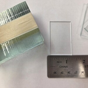 May include: A stack of clear glass slides next to a single rectangular glass slide positioned above a metal ruler. The ruler shows the width of the single glass slide is approximately 1 inch. The glass pieces are transparent and rectangular.