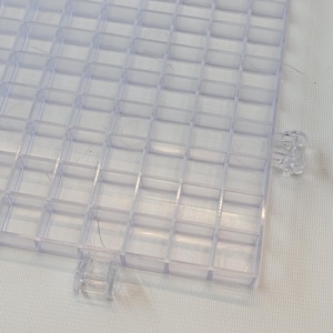 May include: A clear plastic grid with square compartments. The grid has two small plastic tabs on the sides.