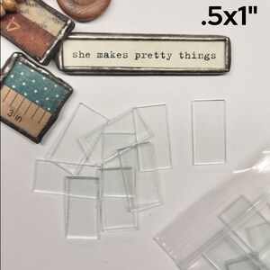 May include: A collection of clear rectangular glass tiles measuring .5x1 inches. Included are finished pieces with metal edges, one featuring the text 'she makes pretty things' and others with patterned paper, alongside loose clear glass rectangles.