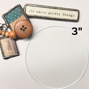 May include: A 3-inch clear glass circle with a wooden button with a cross design and a small framed piece of fabric with a white and blue pattern. The text "she makes pretty things" is visible on a silver framed piece of paper.