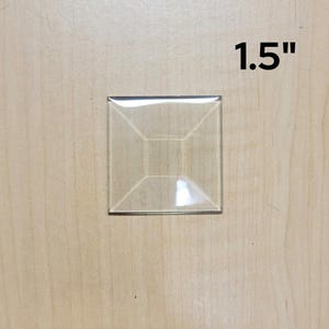 May include: A clear, square glass tile, measuring 1.5 inches across, is centered on a light wood surface. The tile has a beveled edge and an interior design that creates a three-dimensional effect.