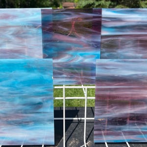 May include: Several pieces of stained glass with a swirling pattern of blue, purple, and white. The glass is arranged on a white grid, with a blurred background of green foliage.