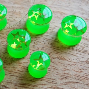 Puede incluir: Seis medidores de oreja de acrílico verde con diseños de estrellas doradas. Los medidores están sobre una superficie de madera.