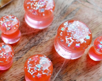 May include: A collection of orange and clear resin ear gauges with embedded silver glitter and star-shaped accents. The plugs are displayed on a wooden surface, with dried seed pods and wisps of grass.