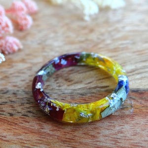 Op de afbeelding: Een kleurrijke harsring met ingebedde gedroogde bloemen en zilveren bladvlokken. De ring heeft een regenboog aan kleuren, waaronder rood, geel, blauw en paars. Geplaatst op een houten achtergrond met roze en witte bloemaccenten.