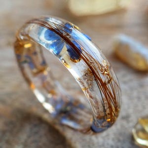 May include: A clear resin ring with embedded blue flowers, strands of brown hair, and gold flakes. The ring is set against a blurred background with other small, light-colored stones. The ring's design is unique and artistic.