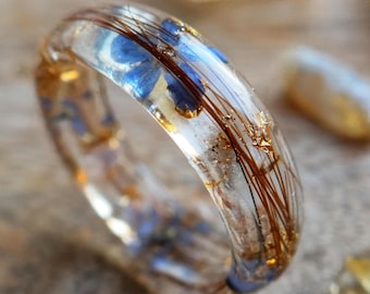 May include: A clear resin ring with embedded blue flowers, strands of brown hair, and gold flakes. The ring is set against a blurred background with other small, light-colored stones. The ring's design is unique and artistic.