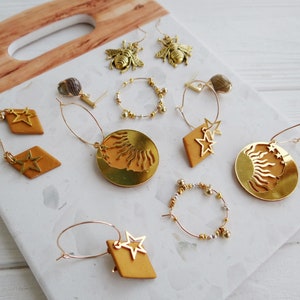 May include: A collection of gold-toned earrings featuring star, sun, and bee designs. The earrings are displayed on a wooden surface with a white background.