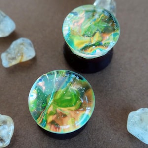 Puede incluir: Dos tapones para orejas de vidrio iridiscente con remolinos verdes y azules. Los tapones están sobre una superficie marrón con varias piedras pequeñas transparentes.