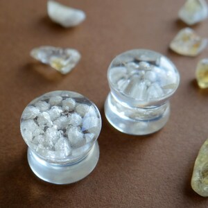 May include: Two clear glass ear gauges with a white crystal design. The gauges are on a brown surface with small pieces of white crystal.